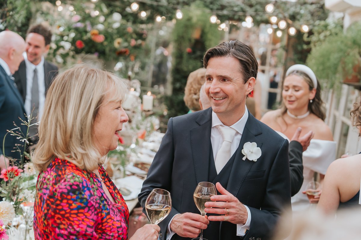  Groom chatting with a wedding guest over drinks, surrounded by flowers and greenery in the Central Greenhouse at Petersham Nurseries, Richmond. 