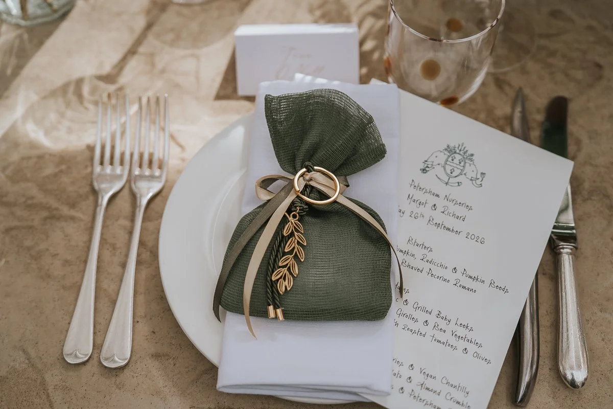  Wedding table setting with a handwritten menu, white napkin and green favour bag in the Central Greenhouse at Petersham Nurseries. 