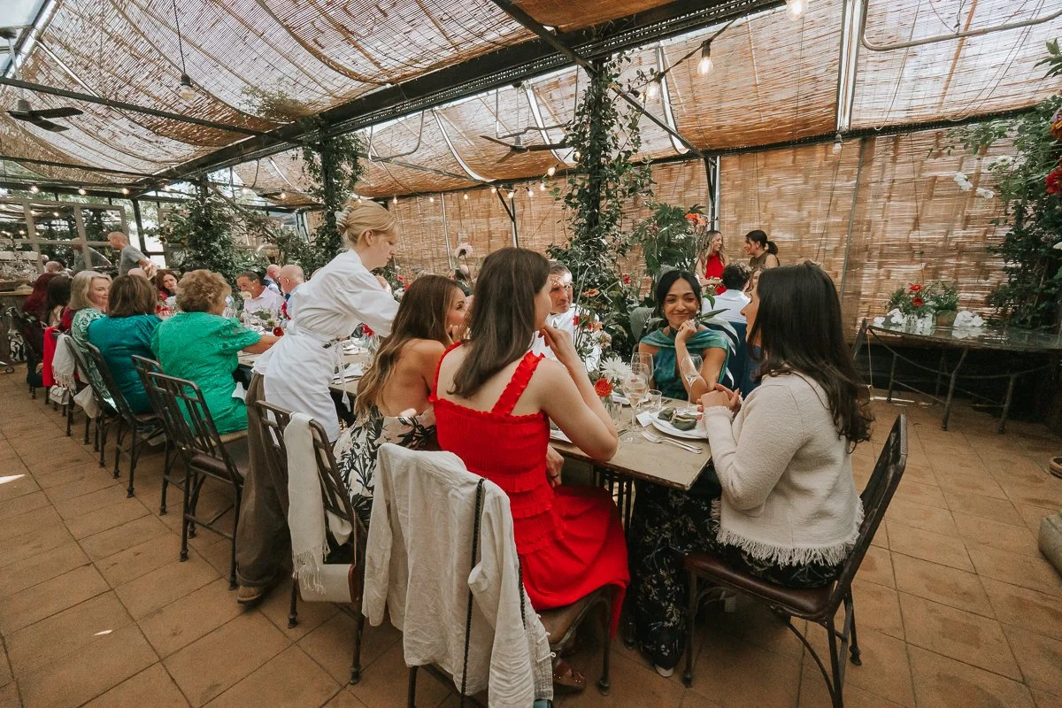  Wedding guests seated at long tables beneath the glass roof, woven blinds and climbing plants of the Central Greenhouse at Petersham Nurseries. 
