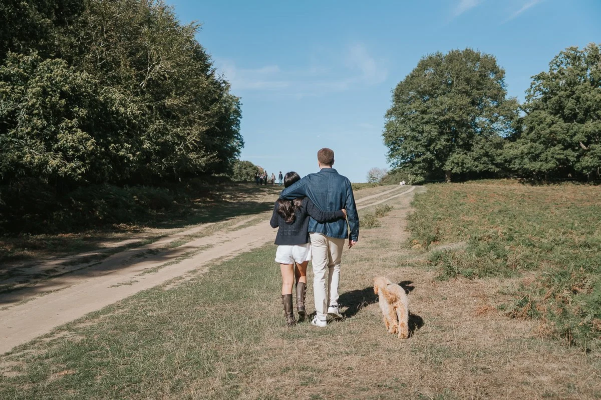  Couple walking arm in arm with their dog beside a tree-lined path during their Richmond Park engagement photoshoot. 