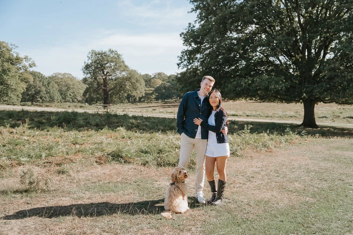  Engaged couple embracing on sunlit grass in Richmond Park, with their dog sitting beside them. 