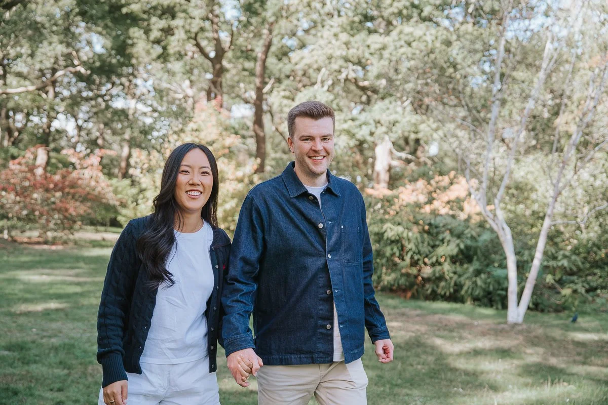  Smiling couple holding hands among leafy trees during their Richmond Park engagement photoshoot. 