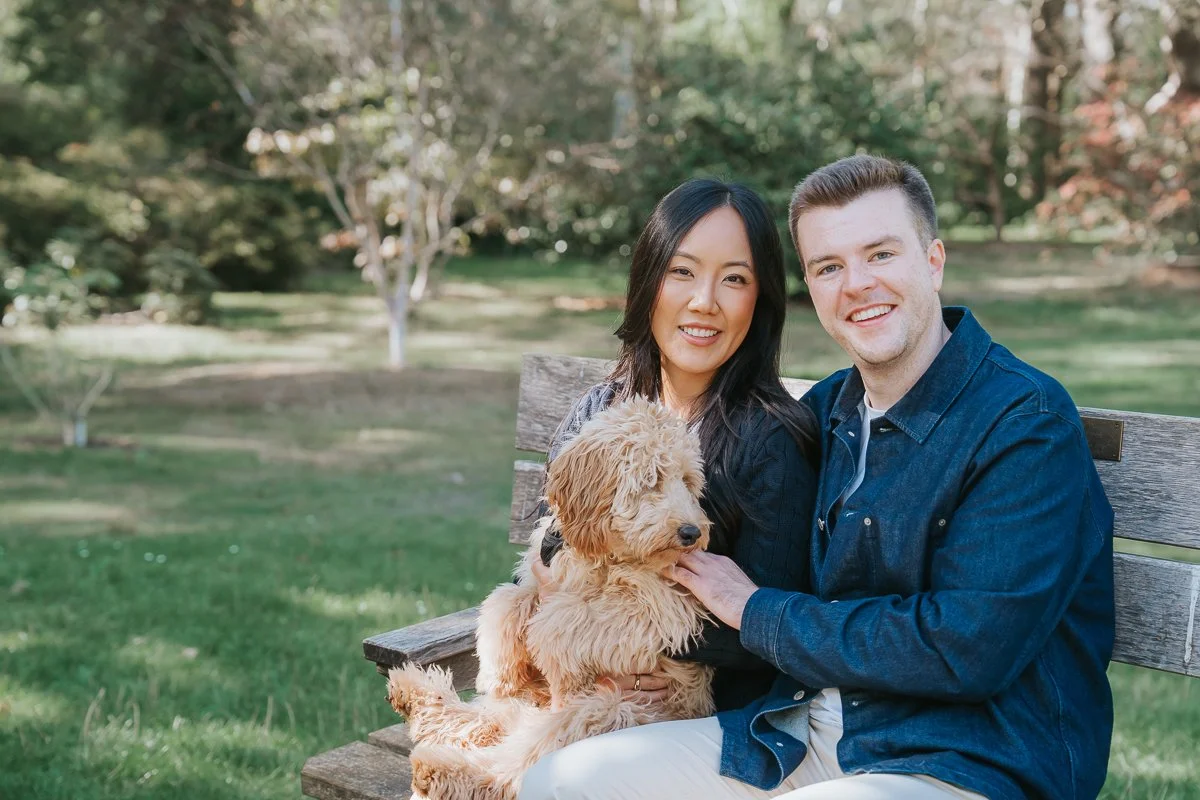  Engaged couple smiling on a wooden bench with their dog on their laps in Isabella Plantation, Richmond Park. 