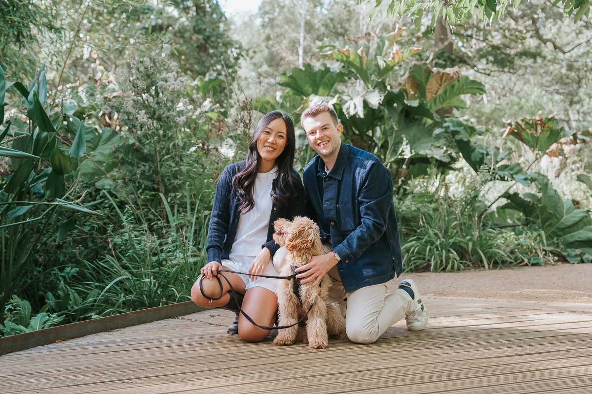  Couple kneeling beside their dog on a wooden boardwalk surrounded by lush foliage during their Isabella Plantation engagement photoshoot. 