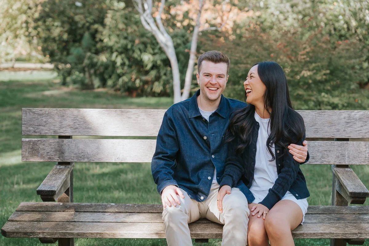  Couple laughing together on a wooden bench beneath leafy trees in Isabella Plantation, Richmond Park. 