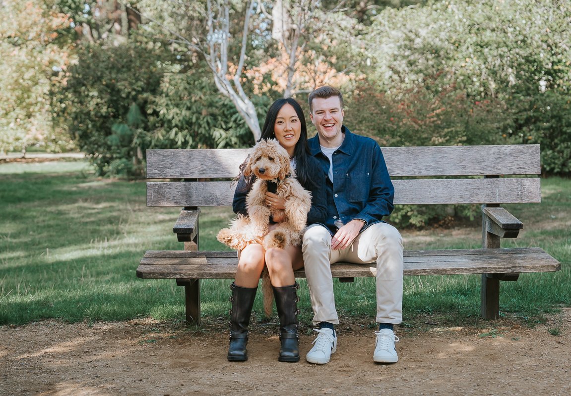  Couple sitting close together with their dog on a garden bench during their engagement photoshoot in Isabella Plantation, Richmond Park. 