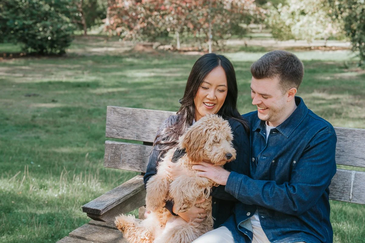  Couple sitting close together with their dog on a garden bench during their engagement photoshoot in Isabella Plantation, Richmond Park. 
