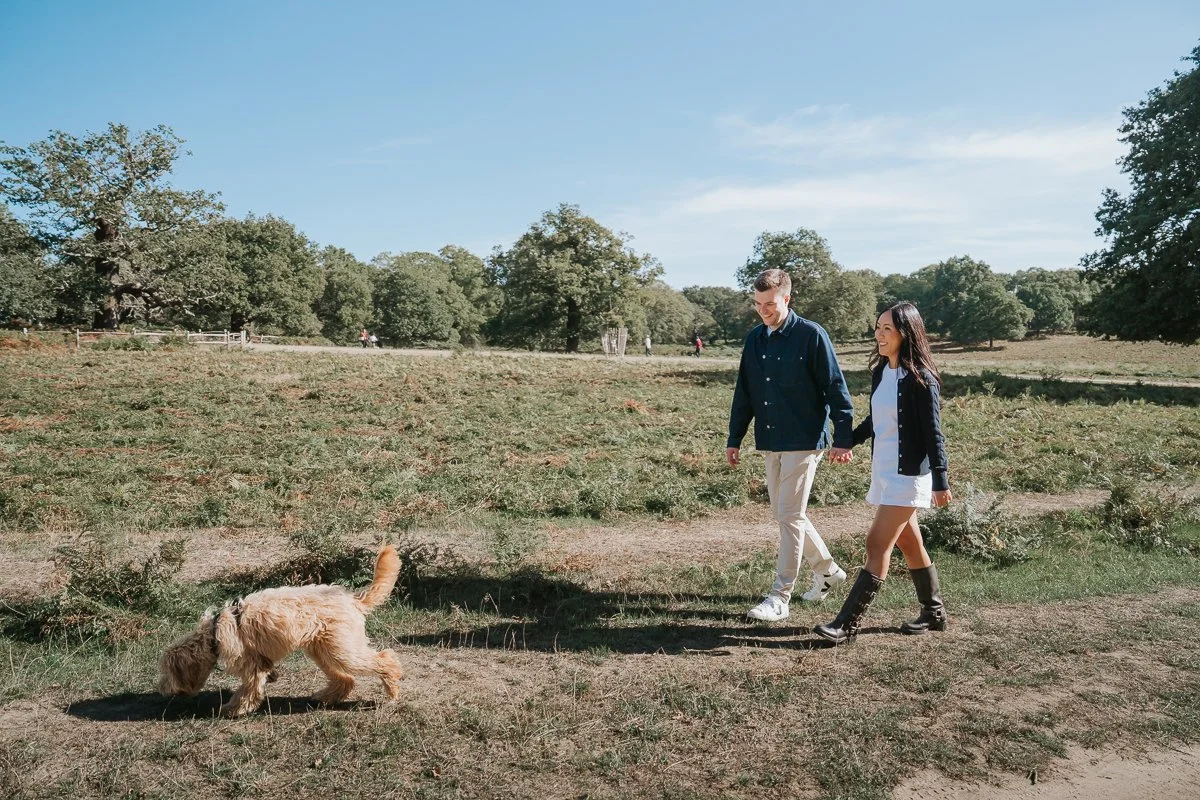  Couple walking arm in arm with their dog beside a tree-lined path during their Richmond Park engagement photoshoot. 