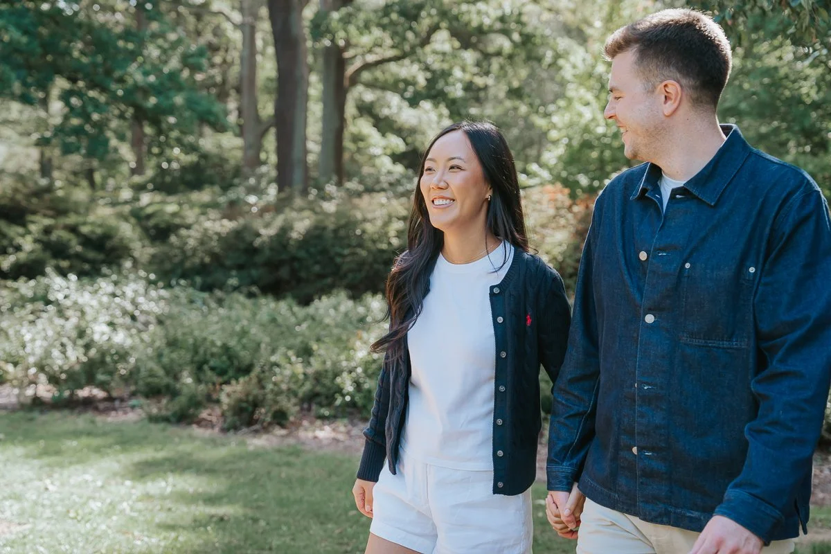  Smiling couple holding hands as they walk past sunlit trees and shrubs in Isabella Plantation, Richmond Park. 