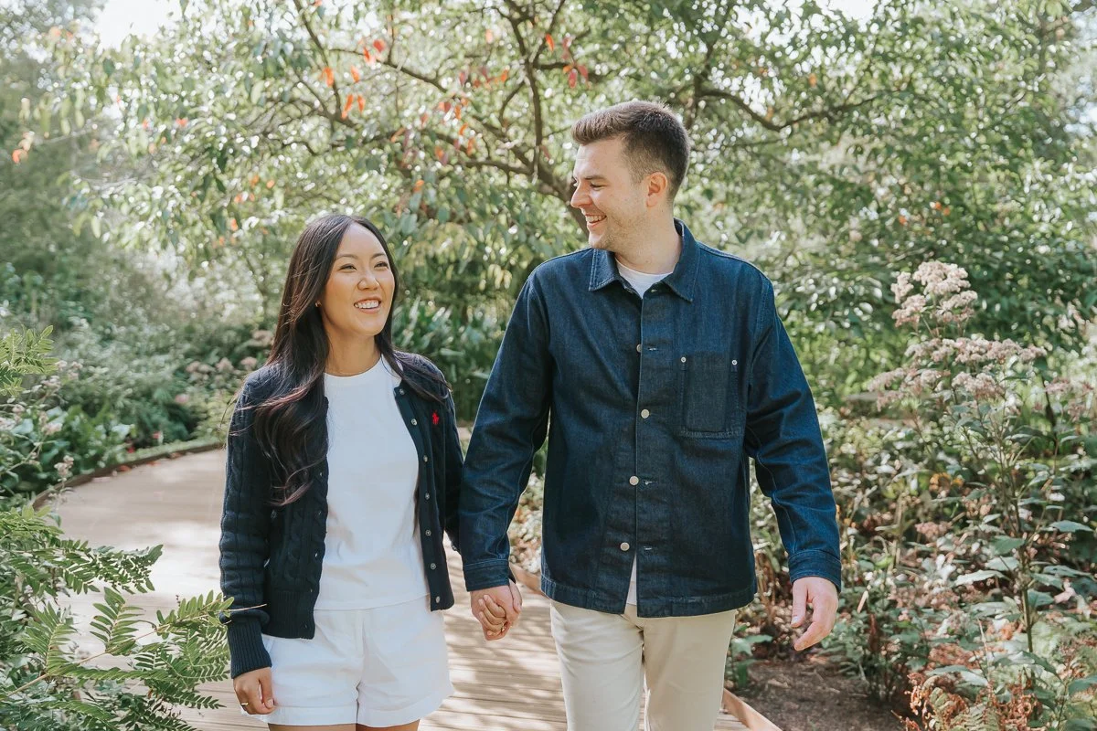  Engaged couple smiling at each other while walking hand in hand along a leafy path in Isabella Plantation, Richmond Park. 