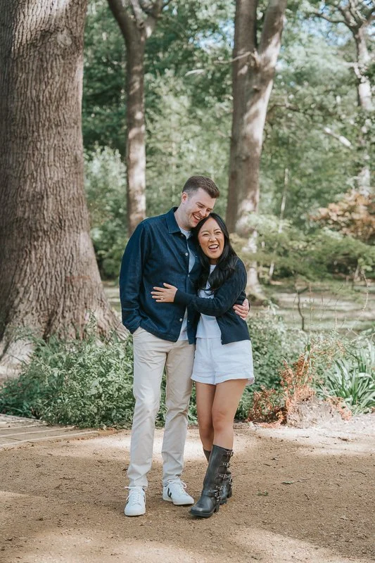  Couple laughing together beneath tall trees during their engagement photoshoot in Isabella Plantation, Richmond Park. 