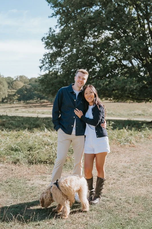  Engaged couple standing arm in arm with their dog on sunlit grass in Richmond Park. 