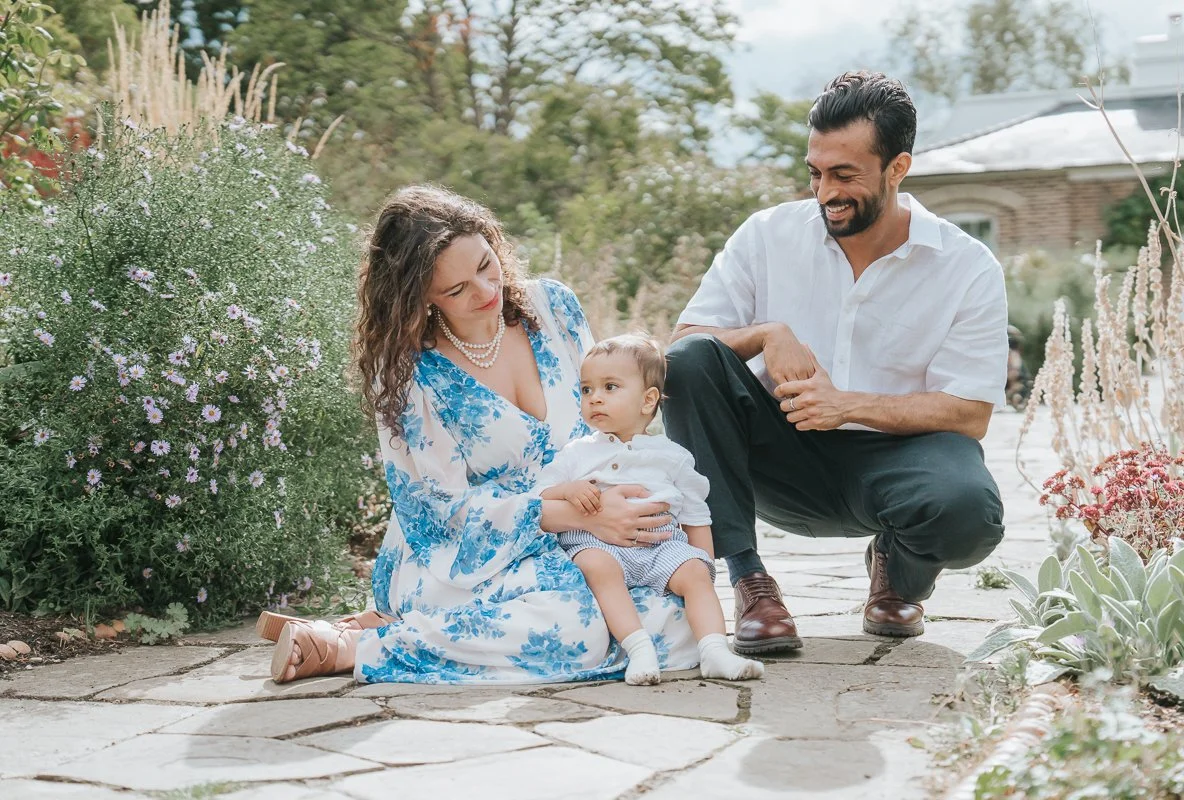  Mother holding her toddler beside smiling dad on a garden path bordered by flowers, photographed by a London family photographer. 