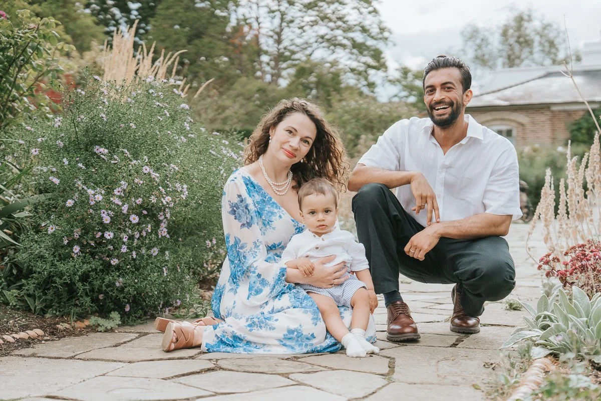  Relaxed family portrait of parents and their toddler sitting together on a stone path surrounded by flowering plants in London. 