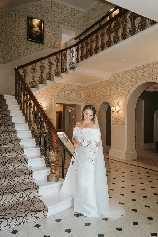  Bride posing next to the elegant staircase at The Petersham Hotel in Richmond, surrounded by ornate Victorian décor and warm natural light, creating a timeless and romantic wedding portrait. 