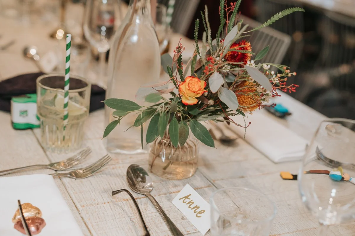  Orange flowers and eucalyptus in a glass vase beside a handwritten wedding place card at The Phene. 