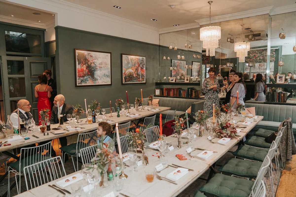  Guests gathering around long wedding breakfast tables beneath chandeliers at The Phene in Chelsea. 