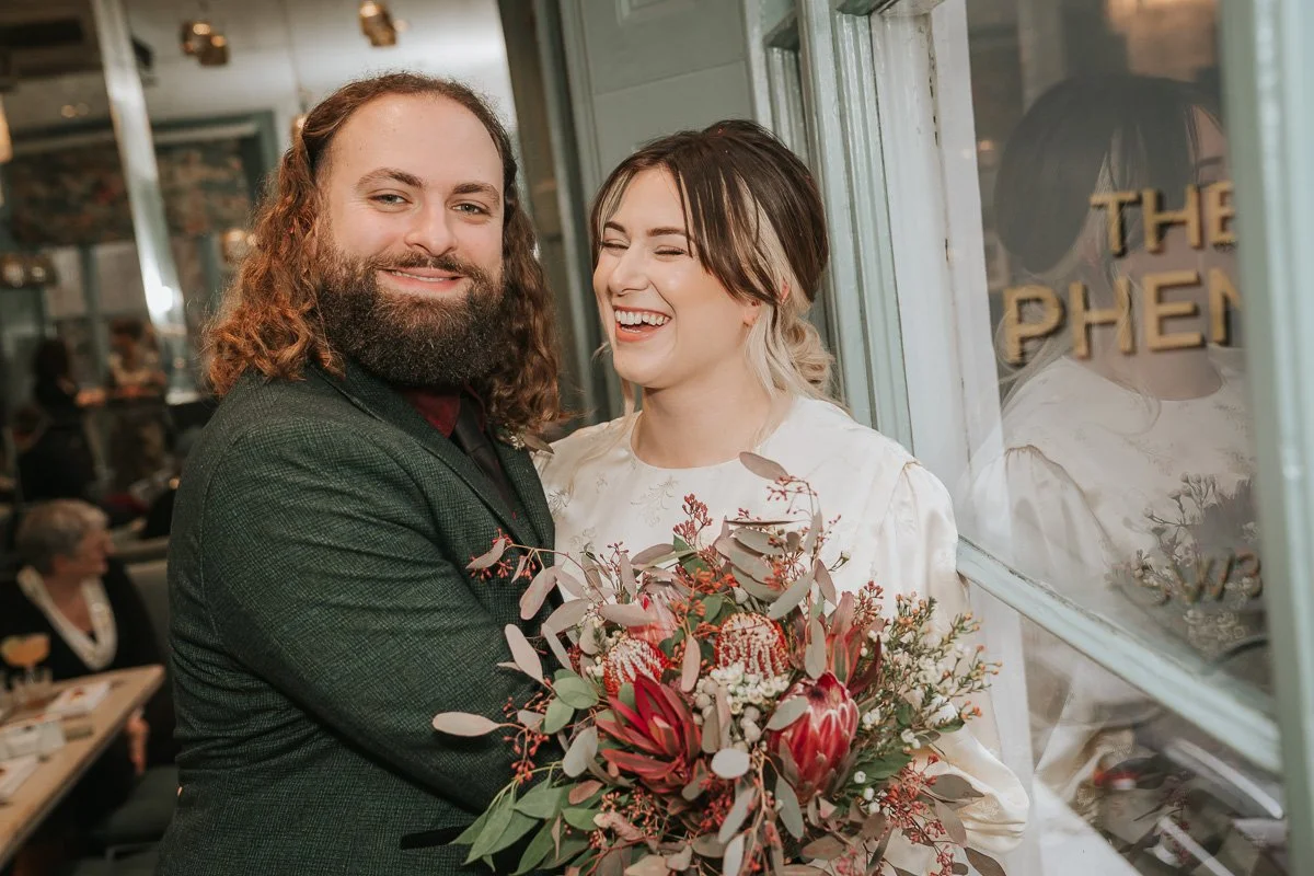  Bride laughing beside the groom, holding a protea and eucalyptus bouquet by The Phene’s window. 