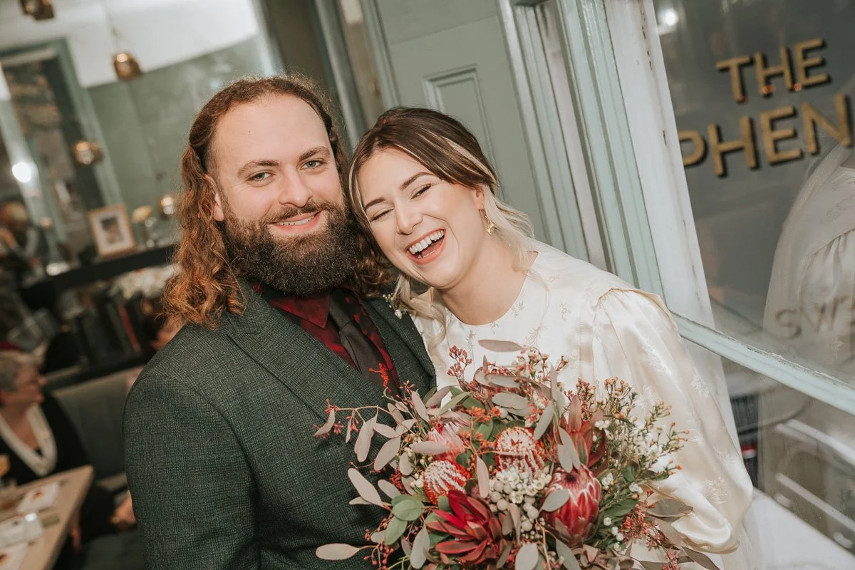  Newlyweds embracing and laughing beside the gold window lettering at The Phene during their Chelsea wedding reception. 