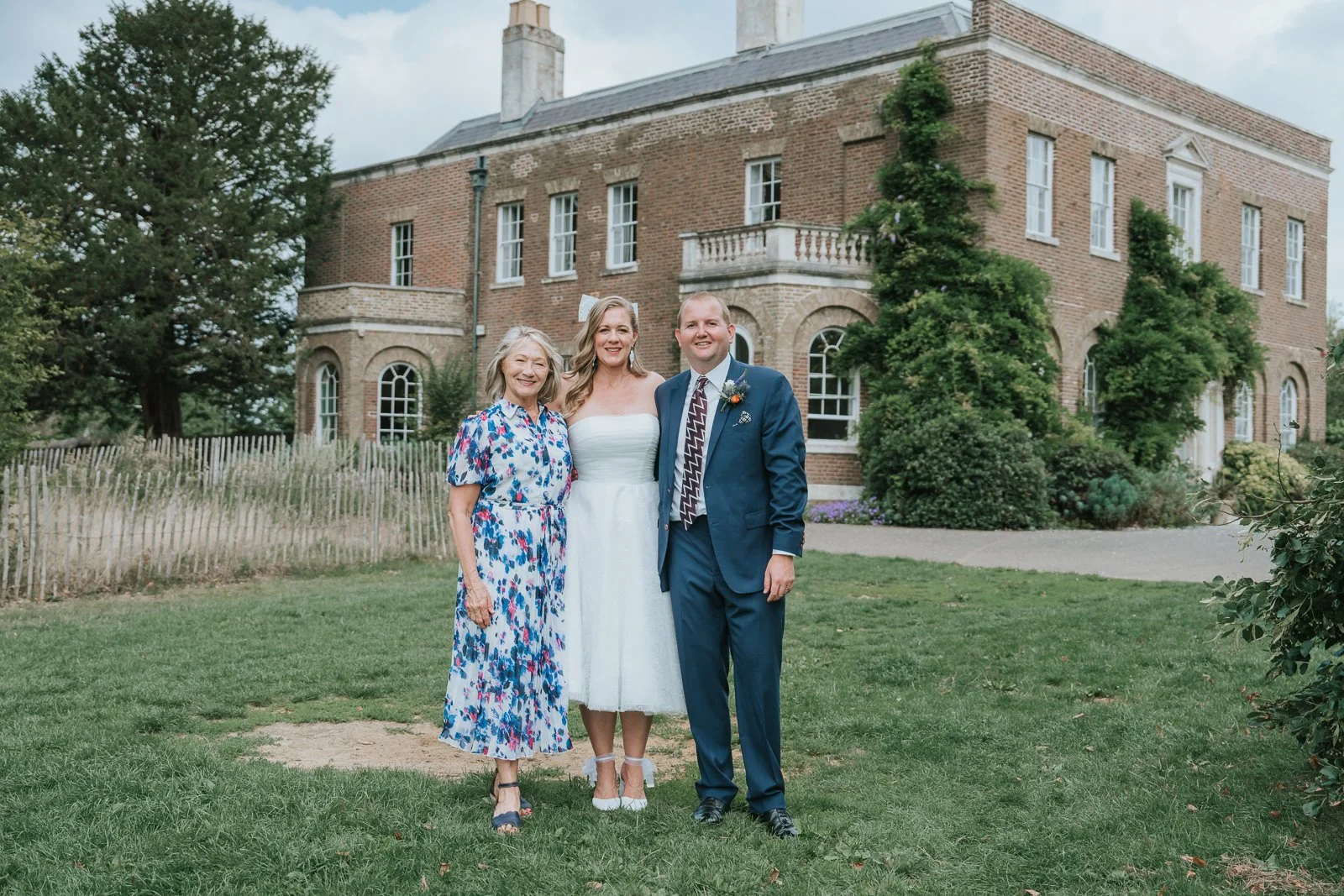  Bride standing between two wedding guests for a group photograph outside Morden Park House. 