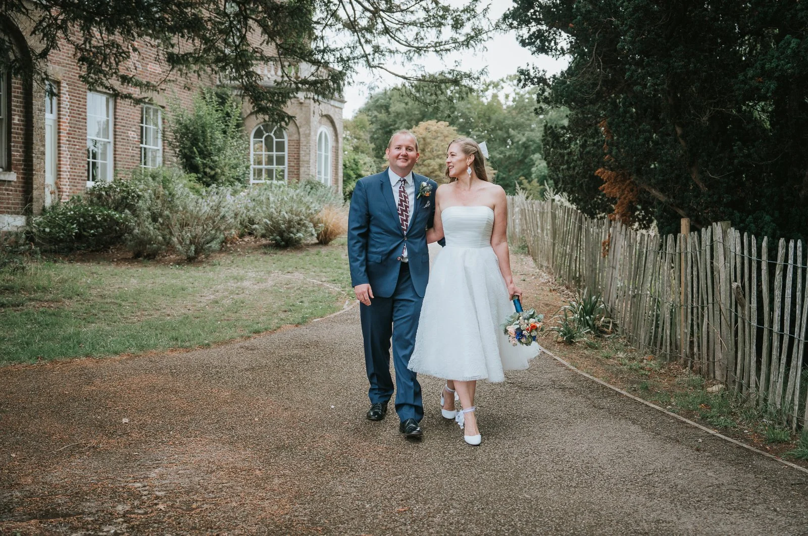  Bride in a tea-length wedding dress walking beside her groom through the grounds of Morden Park House. 