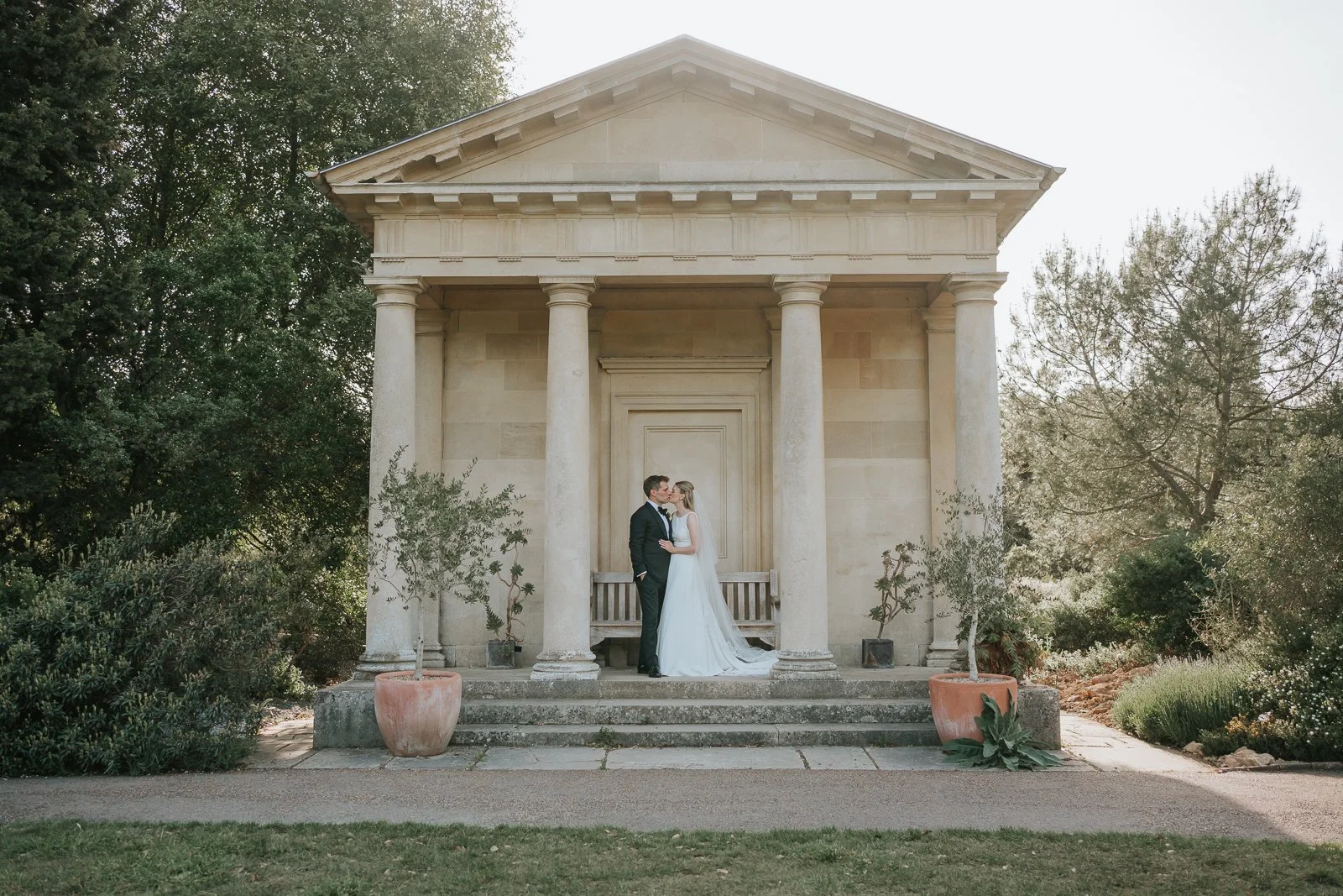 Bride and groom sharing a kiss at the King William Temple in Kew Gardens, framed by classical columns and surrounded by the beautiful landscaped gardens, creating a timeless and romantic wedding portrait.