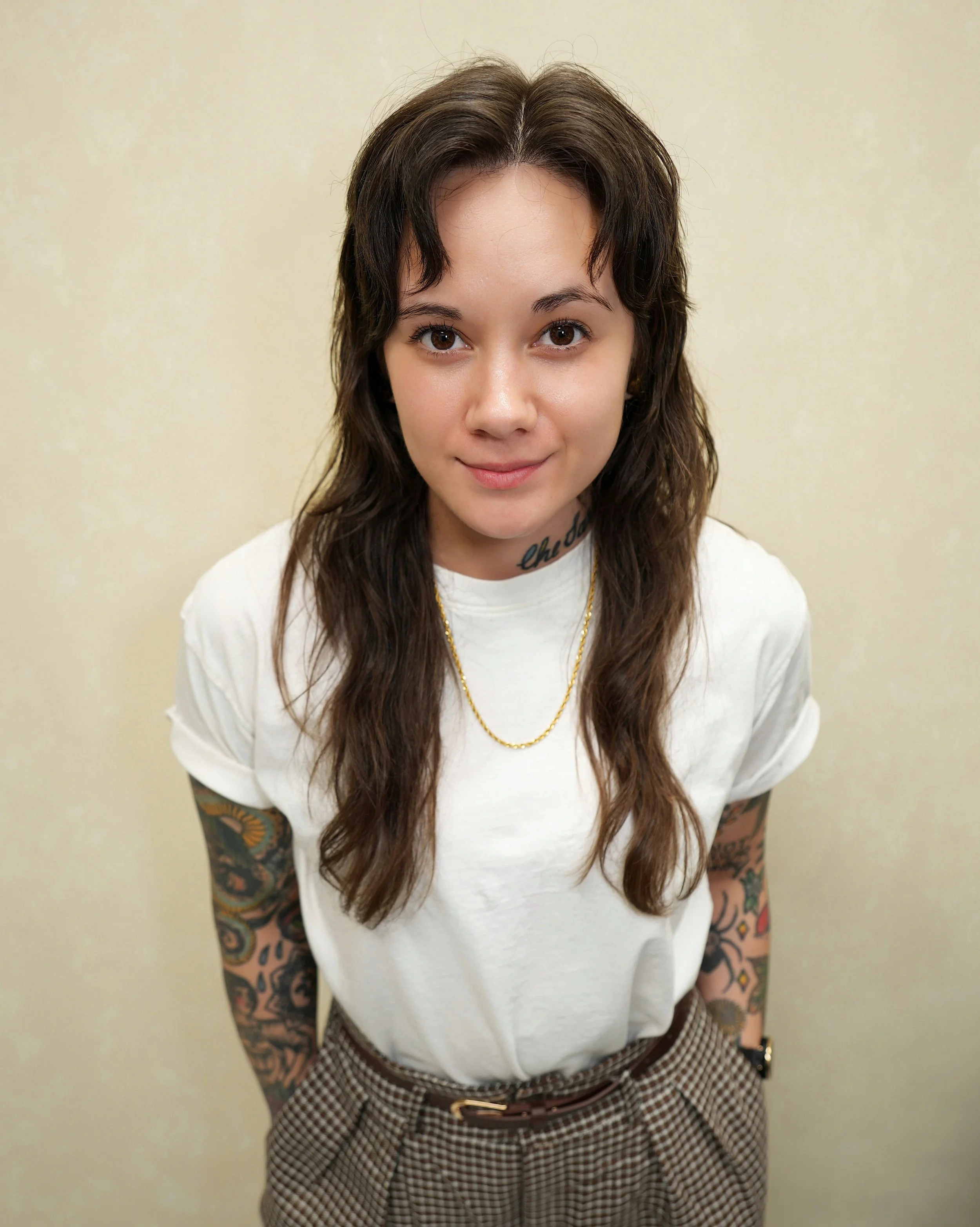 Portrait of a young woman with long wavy brown hair, tattoos on her arms, wearing a white t-shirt, plaid pants, gold chain, and a watch, standing against a plain beige background.