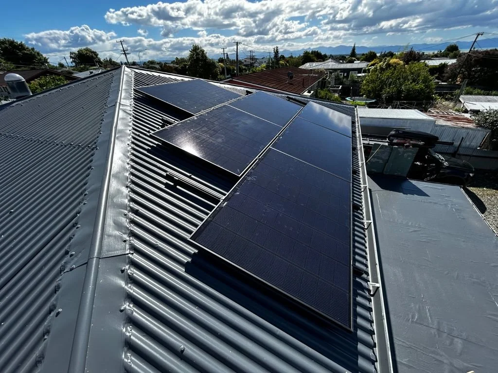 Solar panels installed on a gray metal roof in a residential neighborhood with power lines and trees under a partly cloudy sky.
