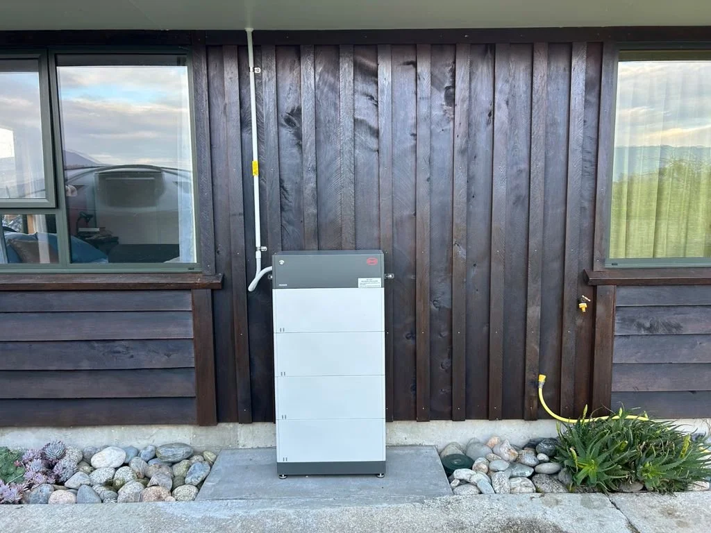 Outdoor electrical box installed on a wooden house wall, with rocks and plants nearby