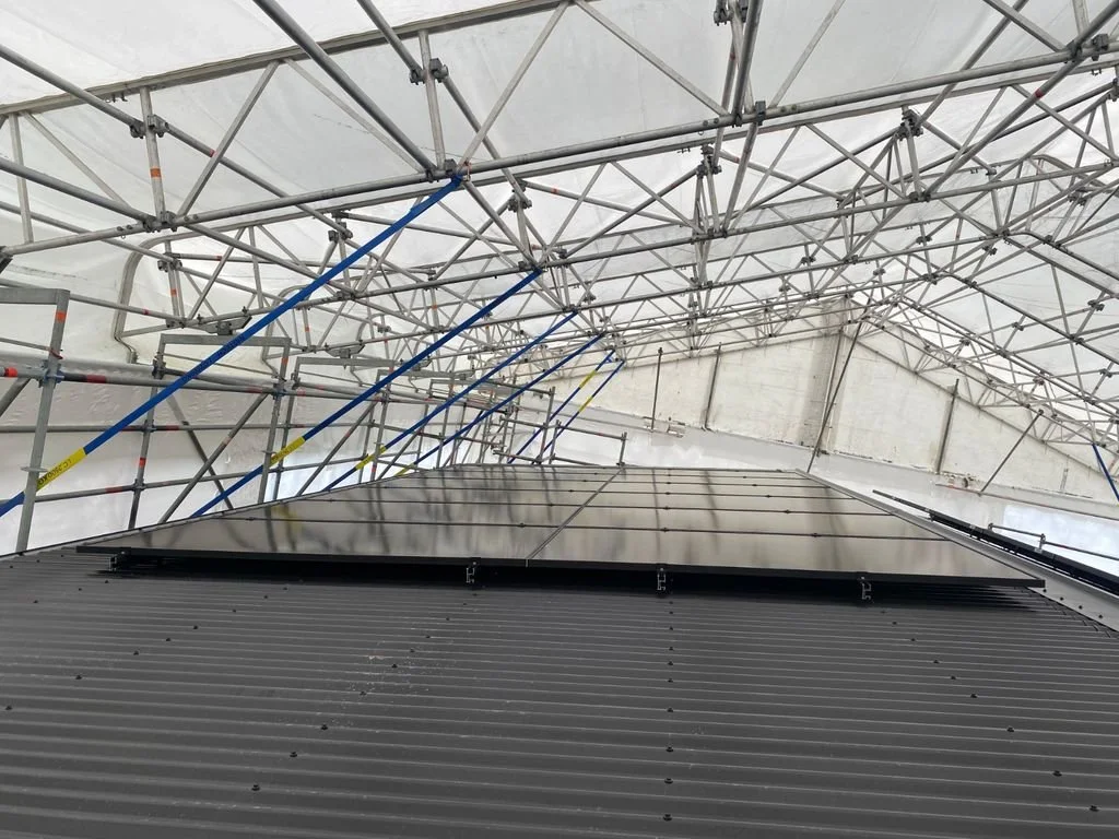 Looking up at a silver solar panel mounted on the side of a building with scaffolding and a white tent-like cover overhead.