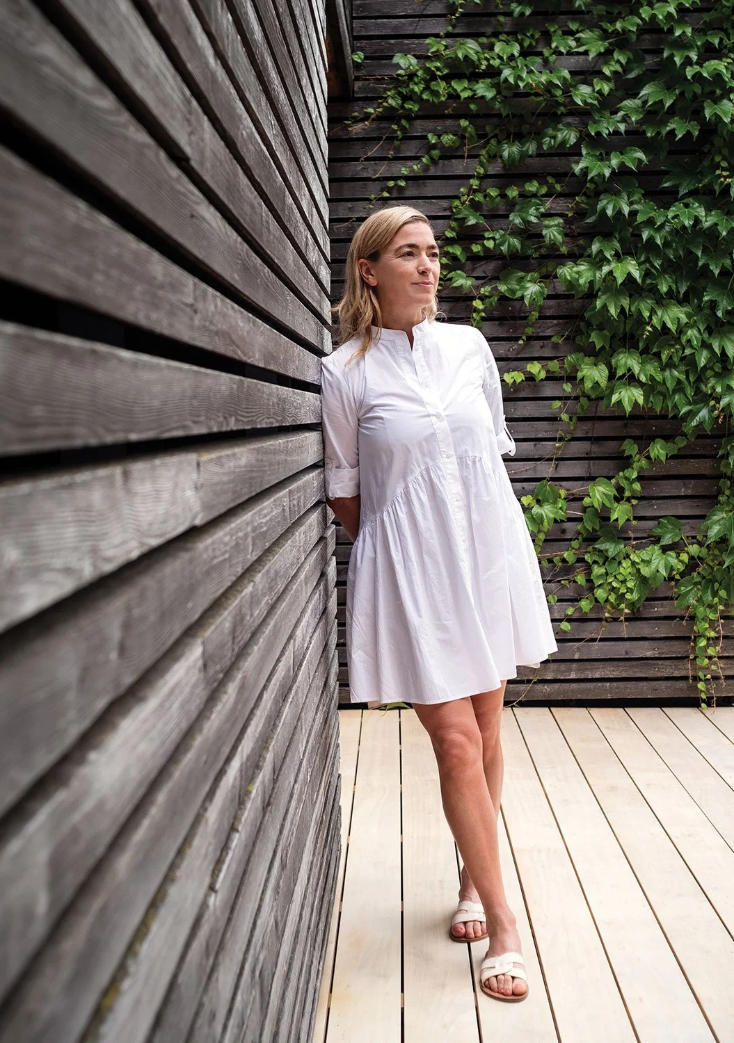 A woman wearing a white dress and sandals standing on a wooden deck with one side against a wooden wall and the other side with green ivy on a wooden fence.