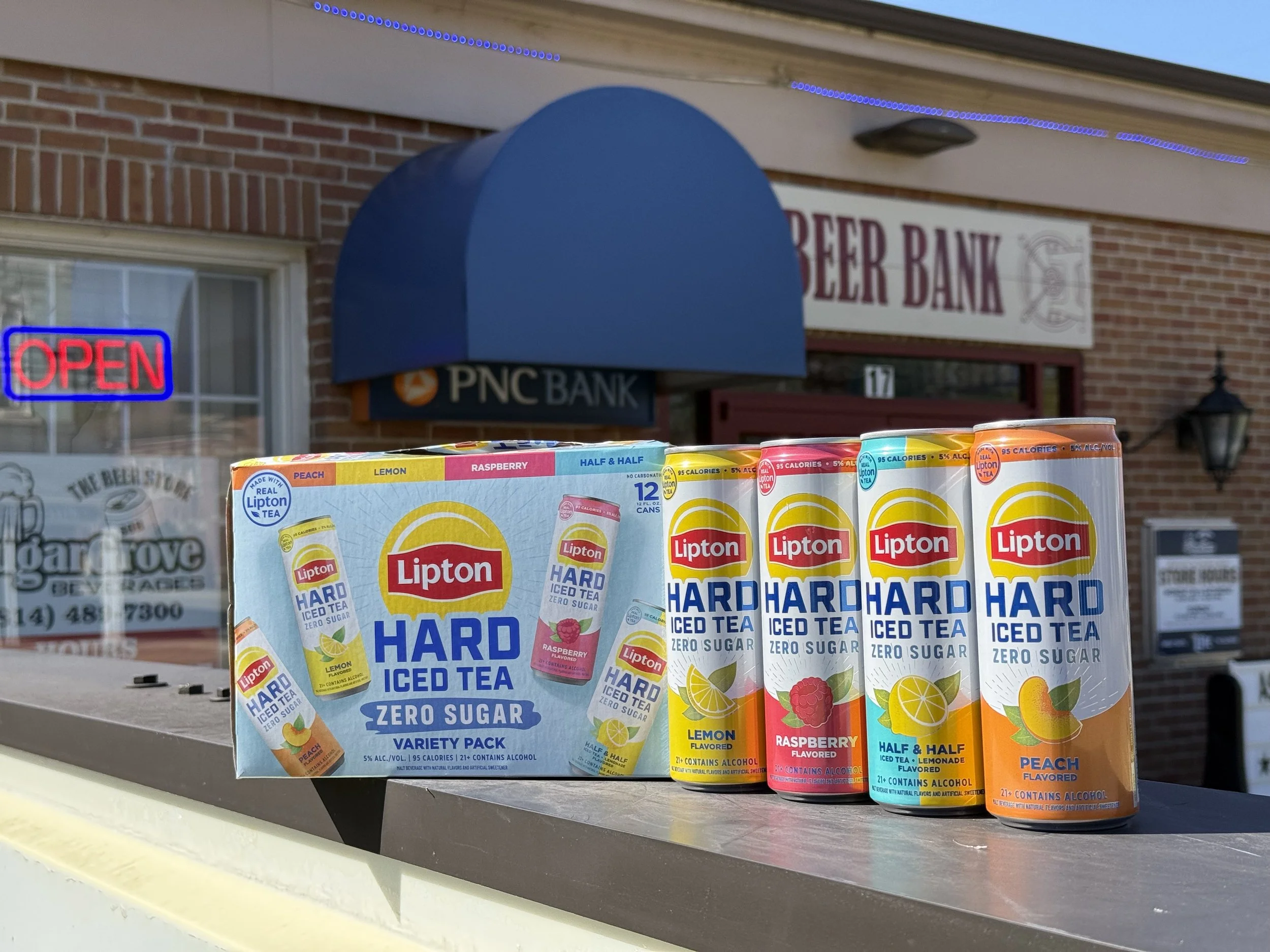 A display of Lipton Iced Tea boxes and cans, including Peach, Lemon, Raspberry, and Half & Half flavors, arranged outside a building with a brick facade and signs indicating a bank and an open sign.