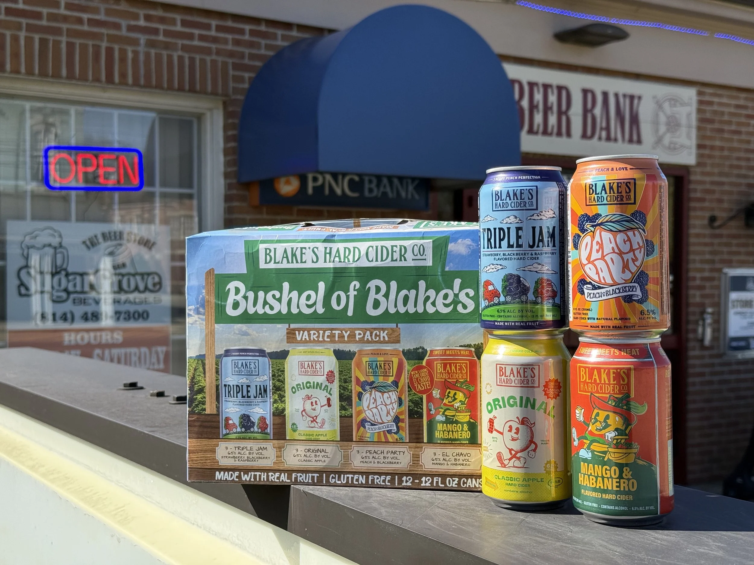 Display of Blake's Hard Cider flavored hard ciders, including Mango & Habanero, Peach & Blackberry, and Original with colorful packaging, in front of a brick building with a sign that reads 'Beer Bank' and a neon 'Open' sign.