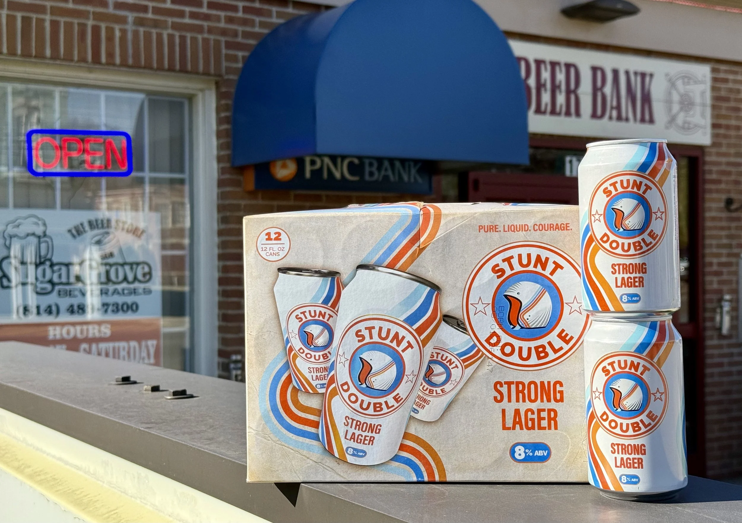 Box of Stunt Double Strong Lager beer cans and can on a sidewalk outside a brick building with an 'Open' sign in the window and a 'Beer Bank' sign above the door.