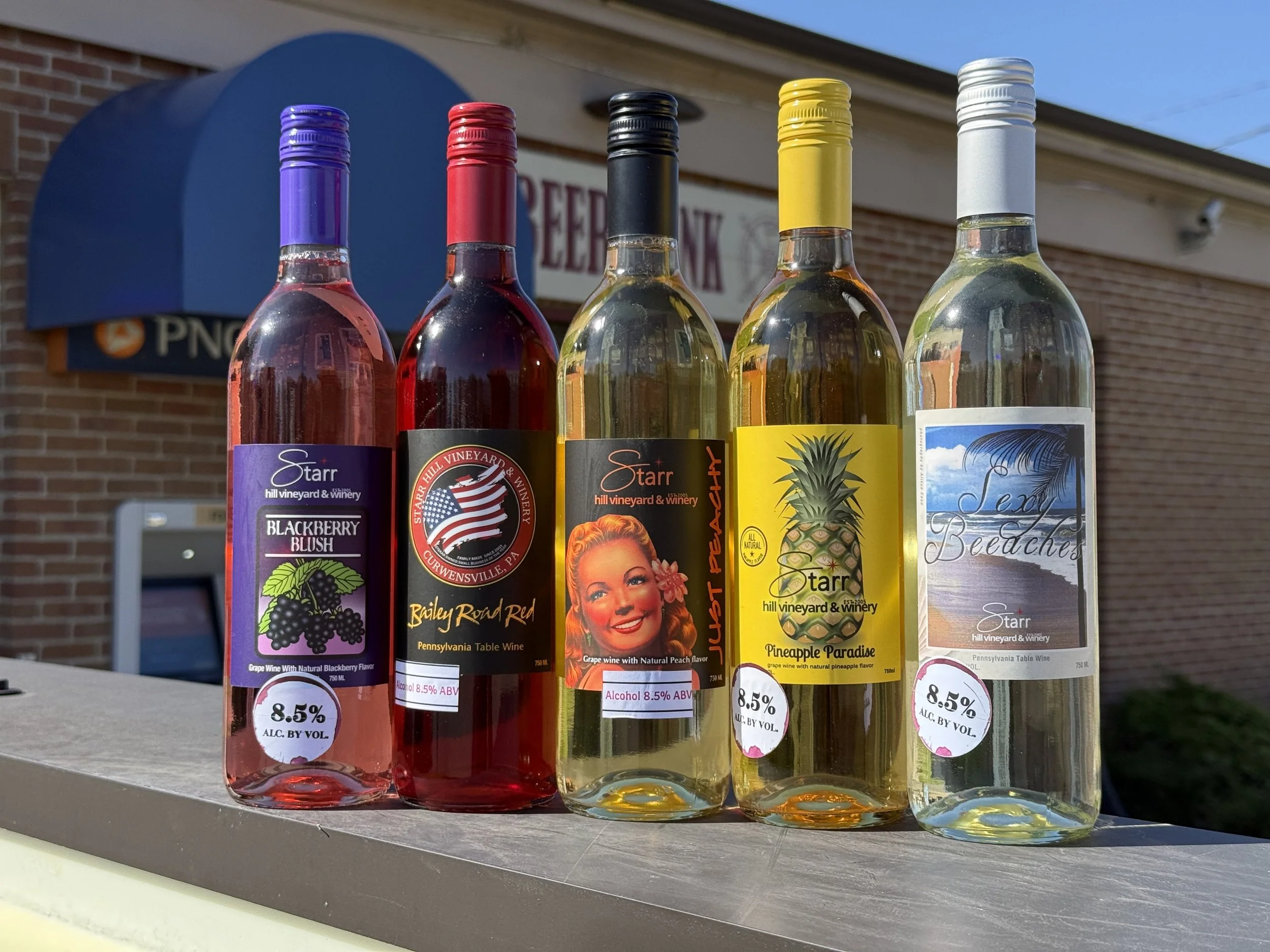 Five bottles of flavored wine displayed outdoors, with a brick building and a sign in the background. The bottles are purple, red, black, yellow, and white and marked with 8.5% alcohol content.
