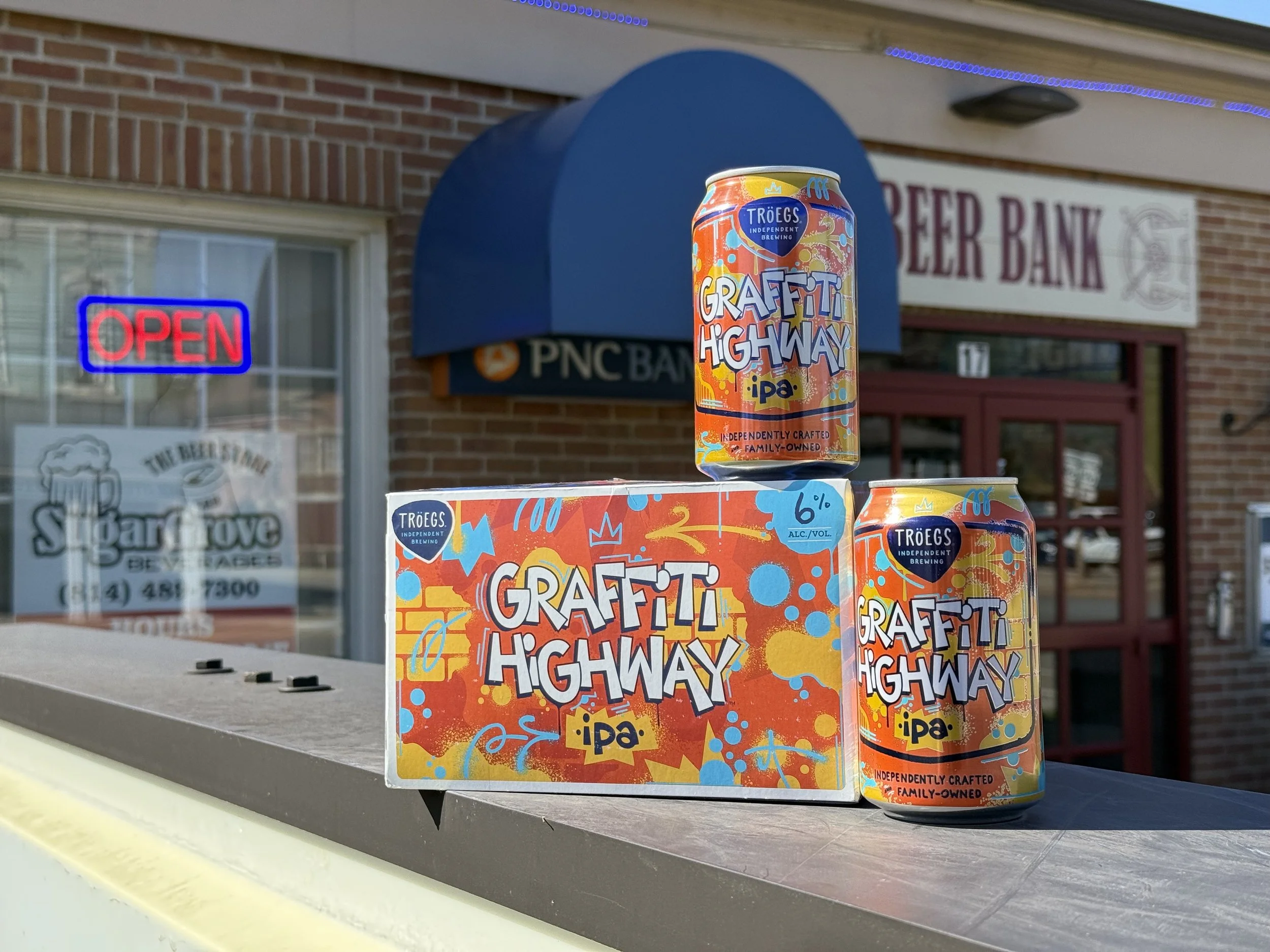 A colorful can of graffiti highway IPA beer on a display box outside a bank building with a blue awning, neon 'OPEN' sign in the window, and a sign reading 'BEER BANK' in the background.