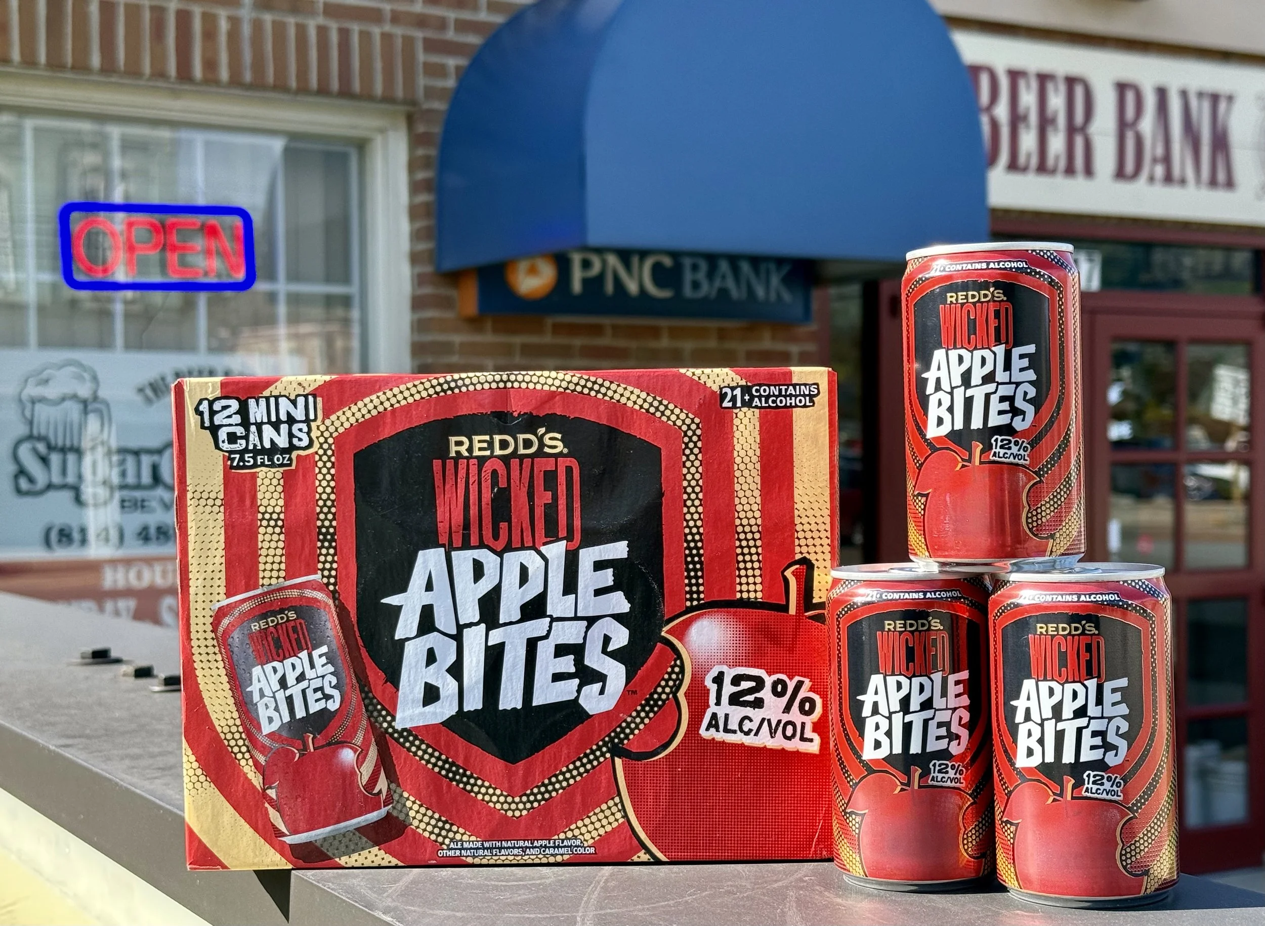 A display of Red's Wicked Apple Bites and cans, with a PNC Bank storefront and an 'Open' sign in the background.