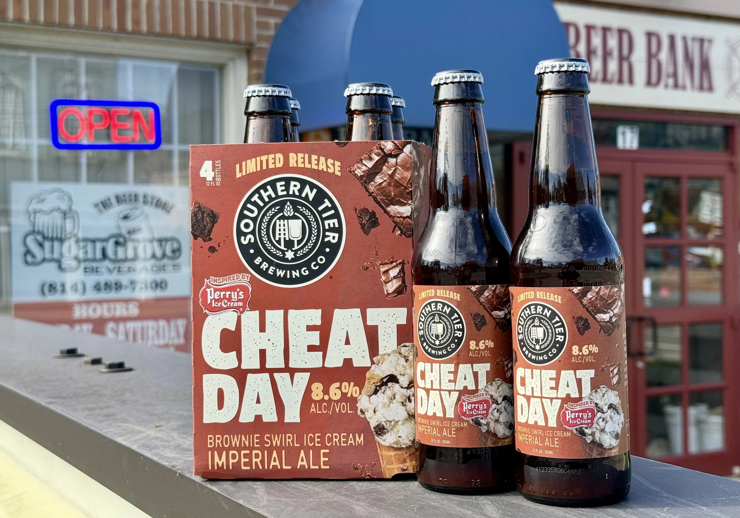 Six bottles of Southern Tier Brewing Co.'s Cheating Day Brownie Swirl Ice Cream Imperial Ale and its packaging on a table outside a store with an 