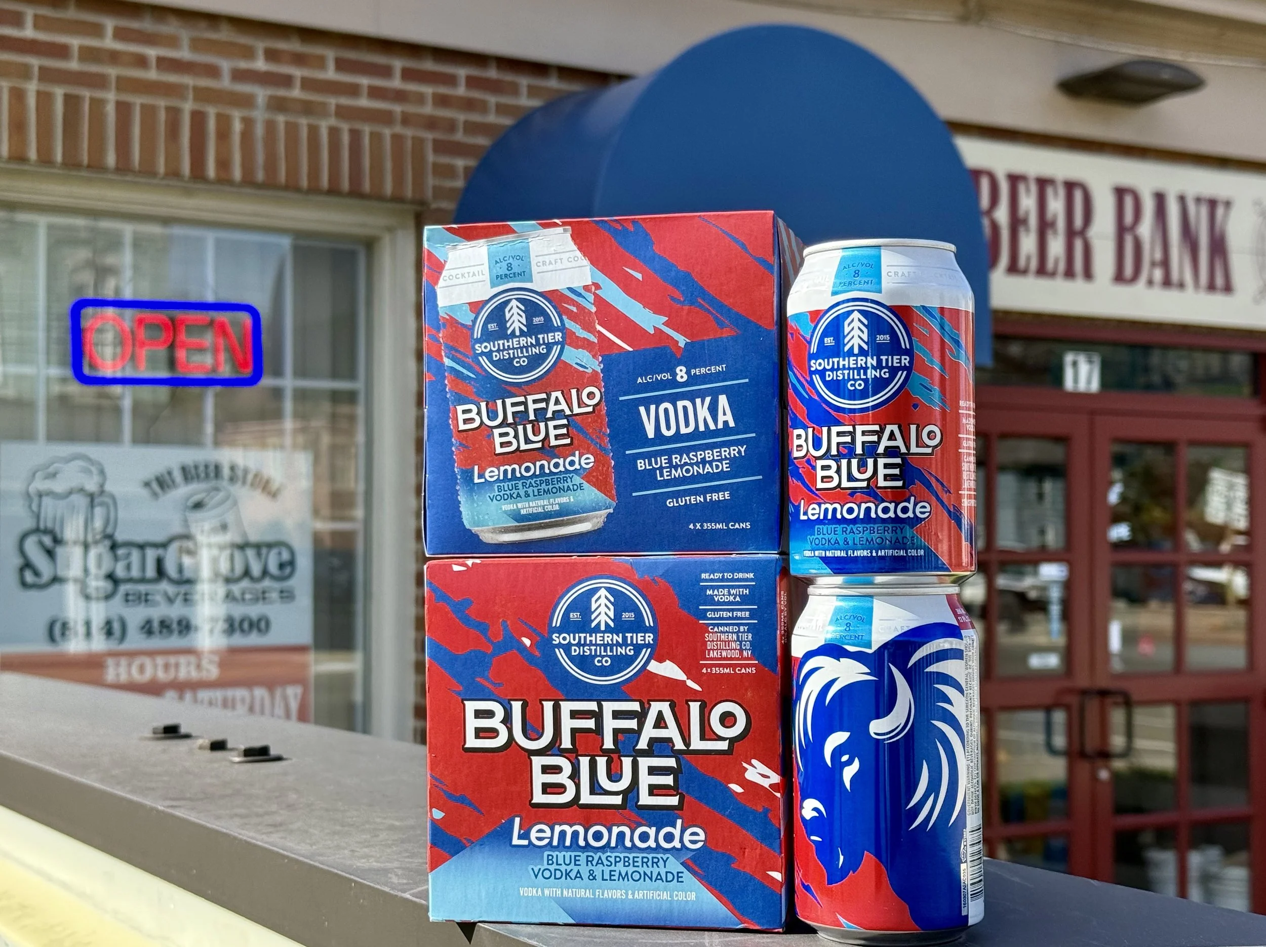 A box and a can of Buffalo Blue Lemonade, a blueberry vodka and lemonade beverage, are displayed outdoors on a table or shelf, with a building and an 'Open' sign in the background.