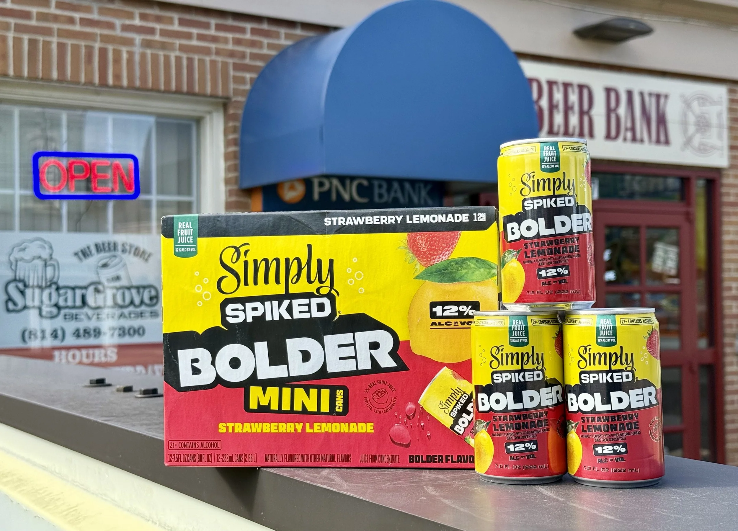 Multiple cans of Simply Spiked Bolder Strawberry Lemonade, a flavored alcohol beverage, stacked on a table outside a store. The background includes a red brick building, a neon 'Open' sign, and a PNC Bank sign.