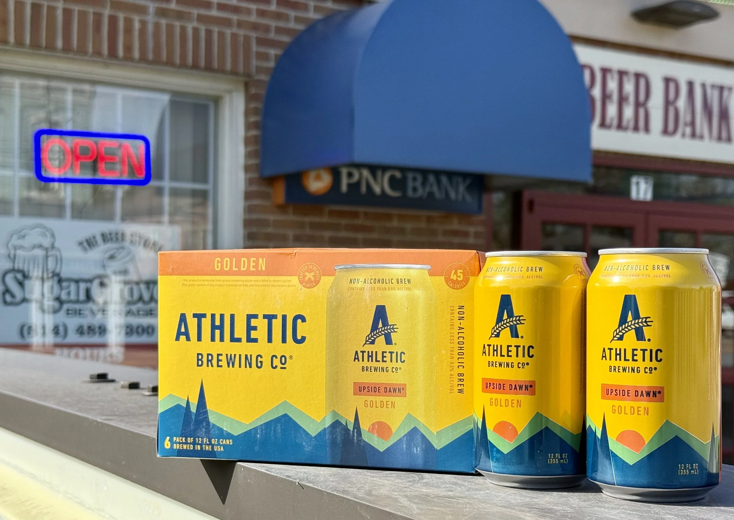 Six yellow cans of Athletic Brewing Co. non-alcoholic beer on a store shelf, with a background of a brick building and a blue bank sign.