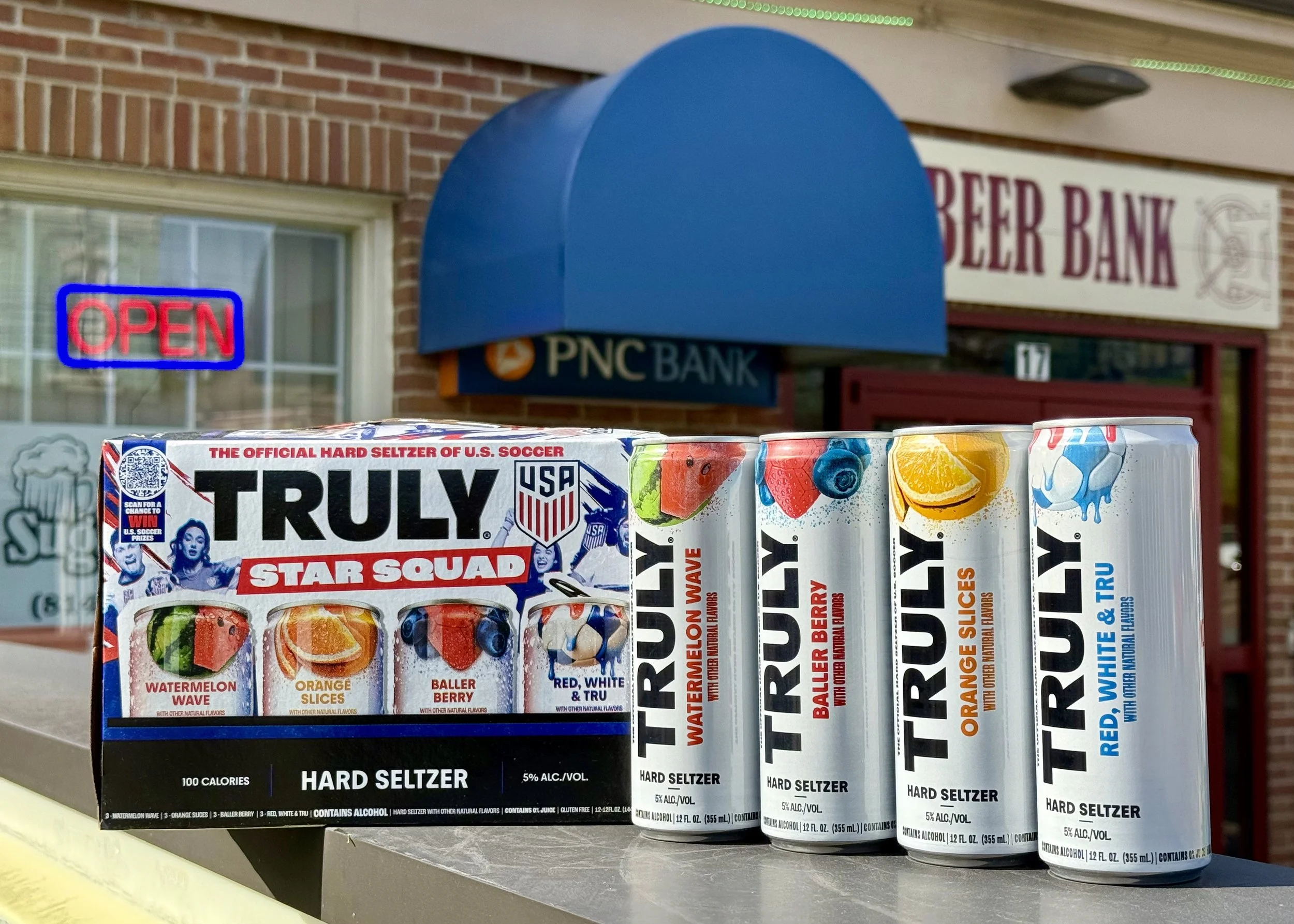 Display of Truly brand hard seltzer cans in front of a PNC Bank branch with an illuminated red and blue 'Open' sign in the window and a blue bank sign outside.