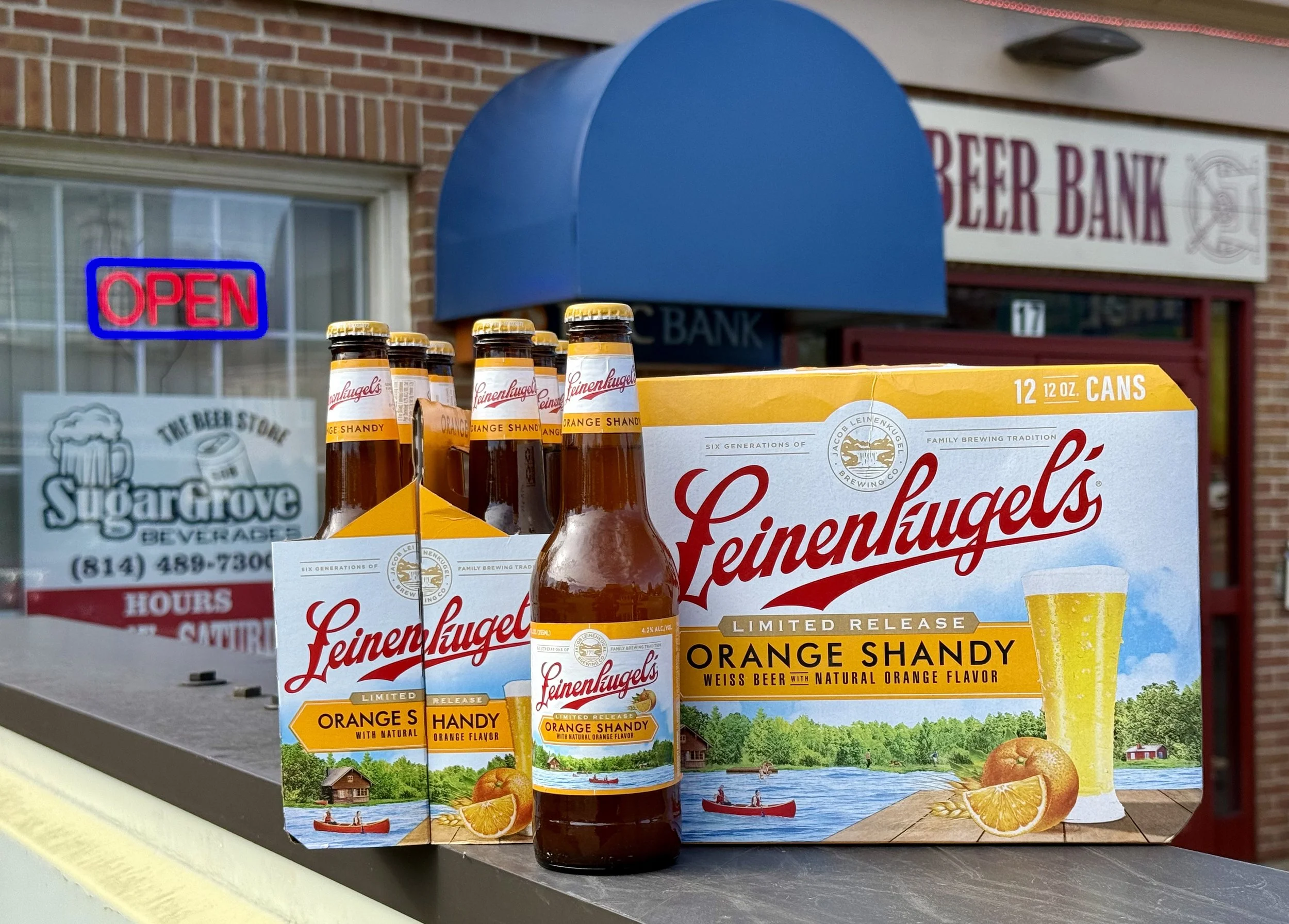 Display of Leinenkugel's Orange Shandy beer bottles and cans outside of a beer store, with a brick wall, a neon 'OPEN' sign, and a sign for Sugar Grove Beverages in the background.