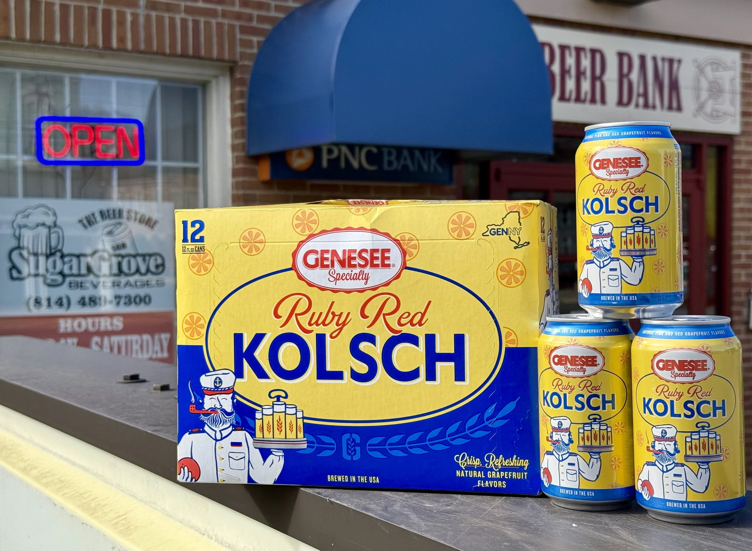 Display of Genesee Ruby Red Kolsch beer cans and a 6-pack, with a PNC Bank sign, a brick building, and an illuminated 'Open' sign in the background.