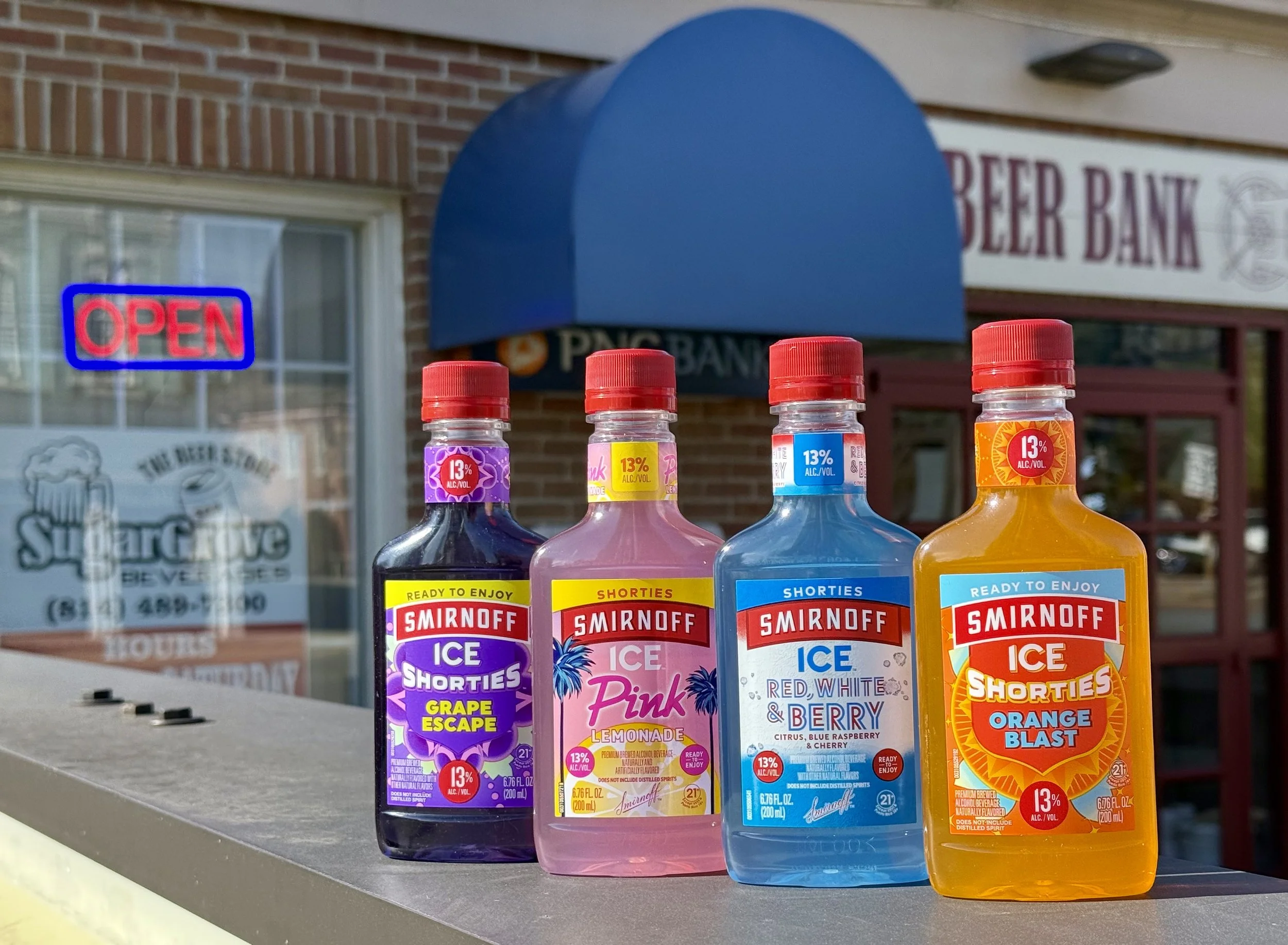 Four bottles of Smirnoff Ice Shorties flavored malt beverages in grape, pink lemonade, citrus, and orange blast varieties, lined up on a bar or table outside a liquor store, with the store's window and signs visible in the background.