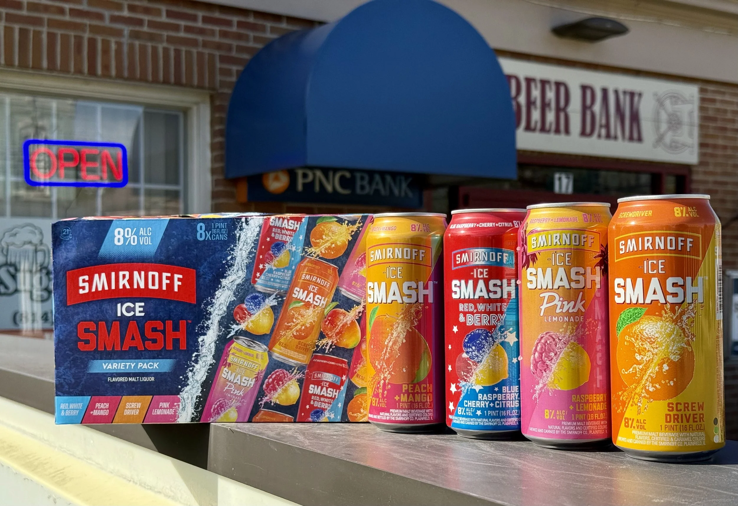 A display of Smirnoff Ice Smash variety pack drinks and a box, with an outdoor store or bank in the background showing a sign for PNC Bank and an illuminated red and blue 'Open' sign.