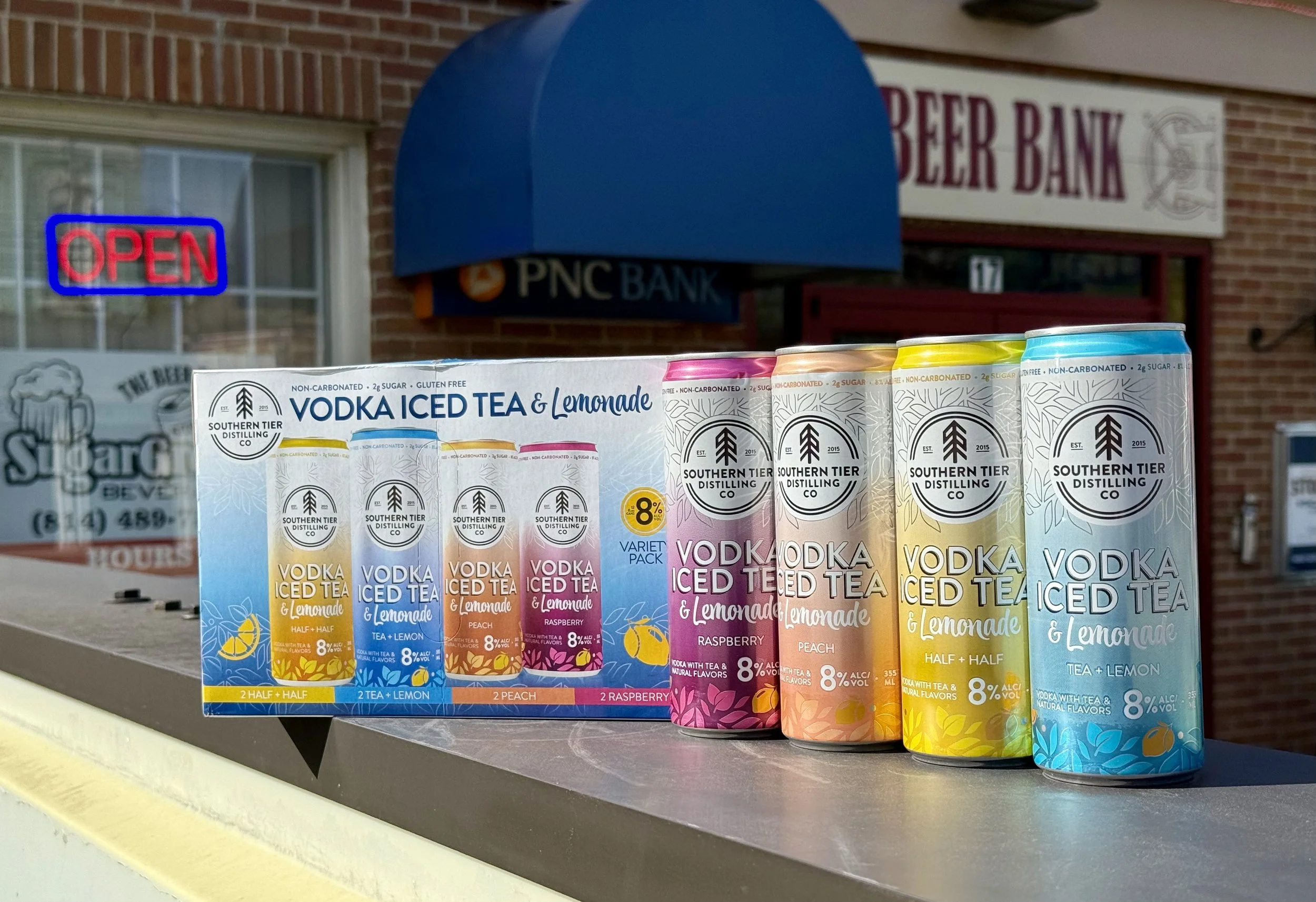 Colorful cans of Southern Tier vodka iced tea and lemonade flavors displayed on a table outside a building with a brick wall, a neon 'Open' sign, and signs for a beer bank and PNC bank in the background.