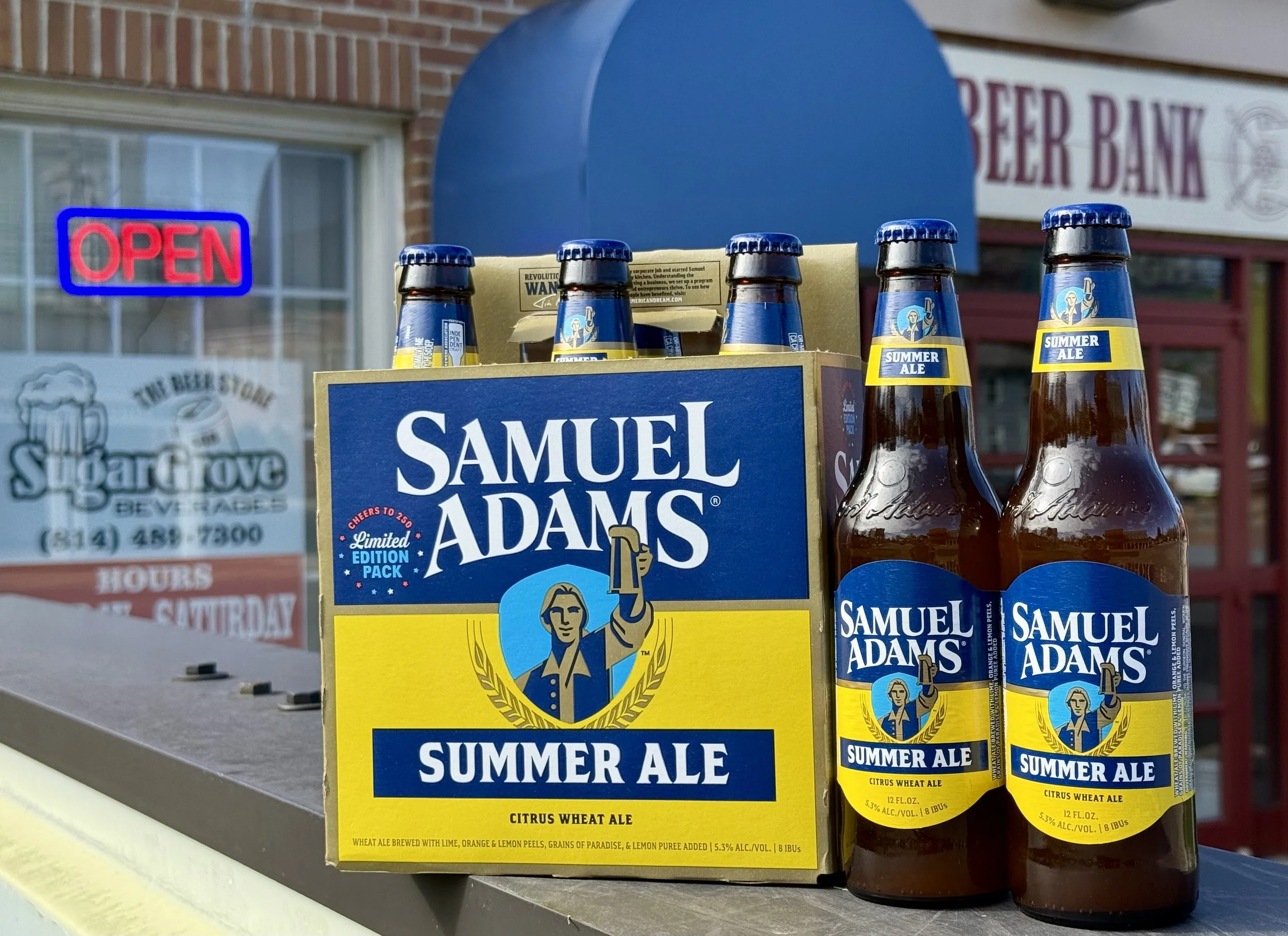 Six bottles of Samuel Adams Summer Ale, a citrus wheat beer, displayed outdoors with a blue and yellow box. In the background, a storefront with an 'OPEN' sign in a window and a sign for 'The Beer Store' is visible.