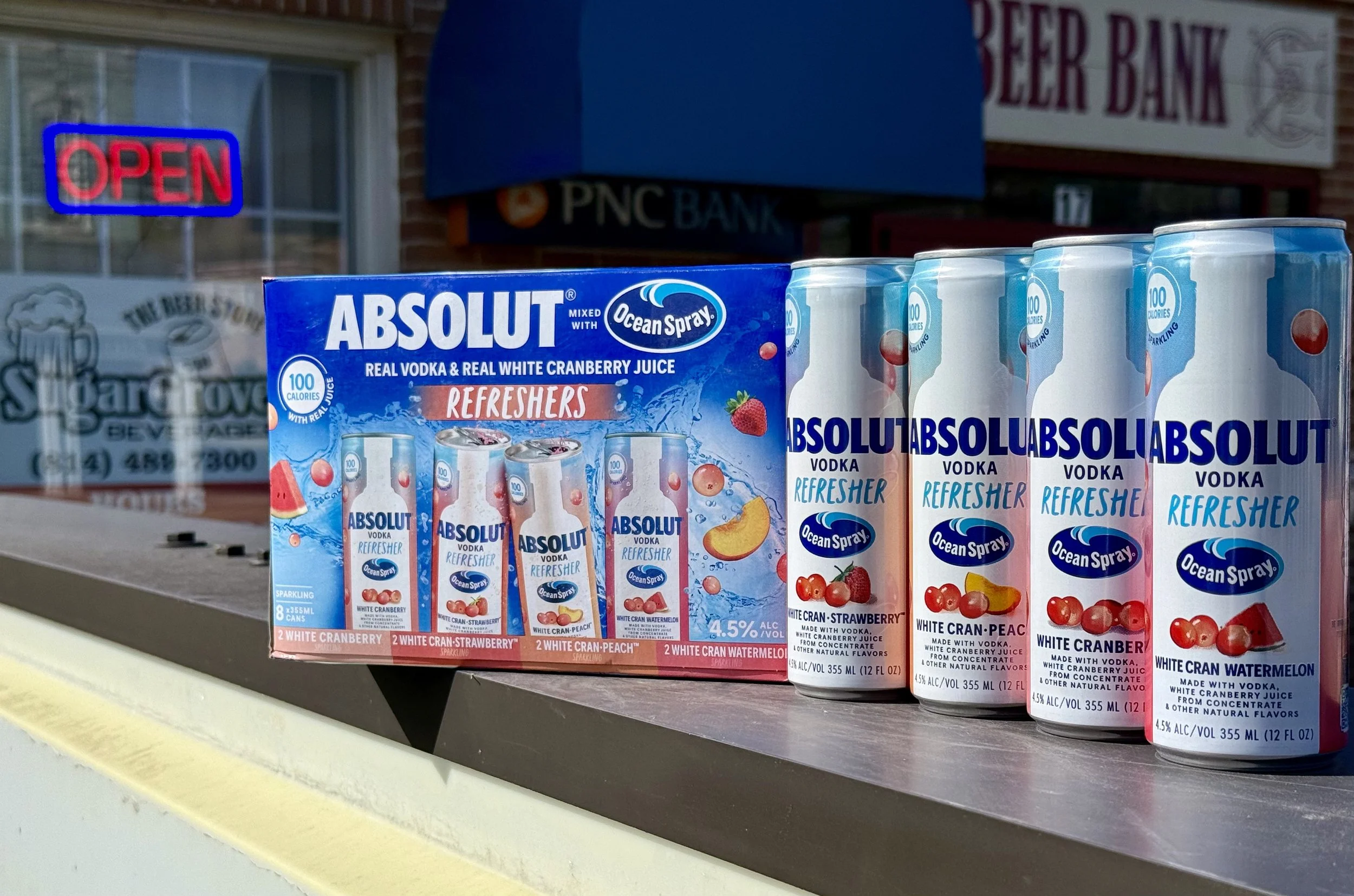 A display of Absolut Vodka beverage cartons with flavor labels, including cranberry, peach, strawberry, and watermelon, on a store shelf outside a bank.
