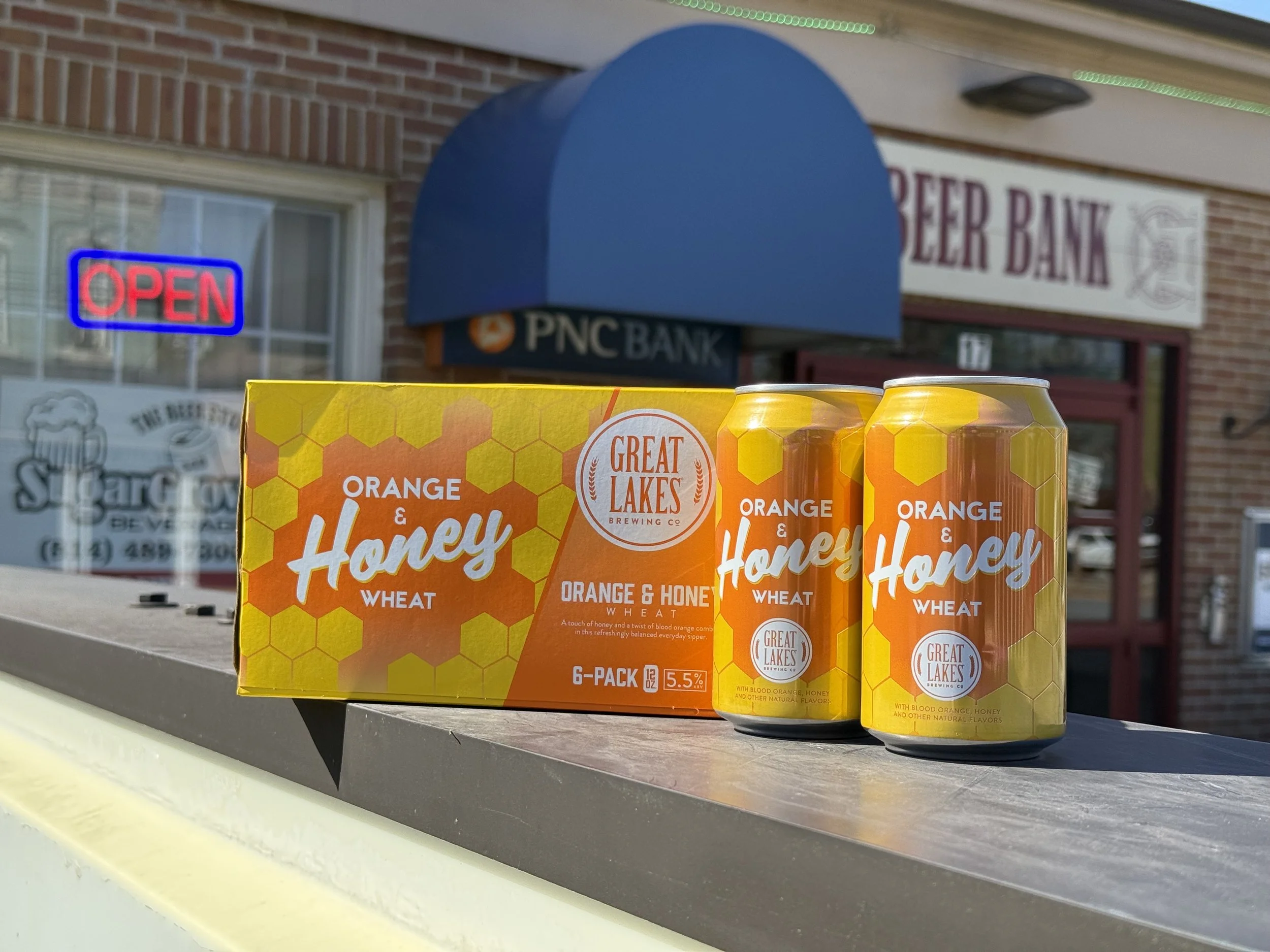 Close-up of a six-pack of Orange & Honey Wheat beer cans from Great Lakes Brewing Co., placed on a ledge outside a building with visible brick wall, window, and shop signs including a neon 'Open' sign and a sign for a beer bank.